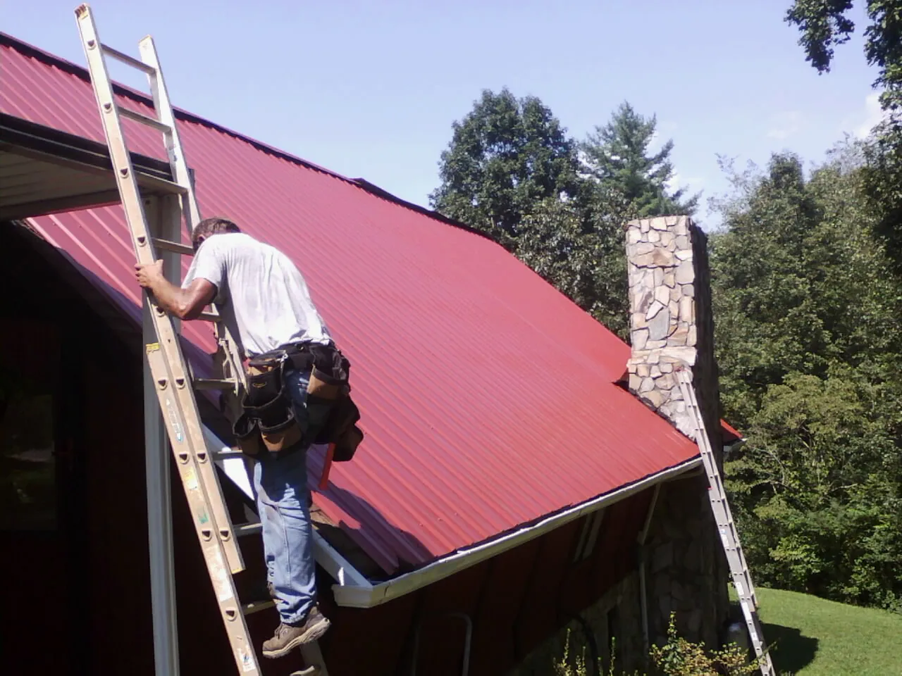 Roofer on a ladder beside a red metal roof and stone chimney — Soffit & Fascia Repair by Roofing Brothers in Mc Dade